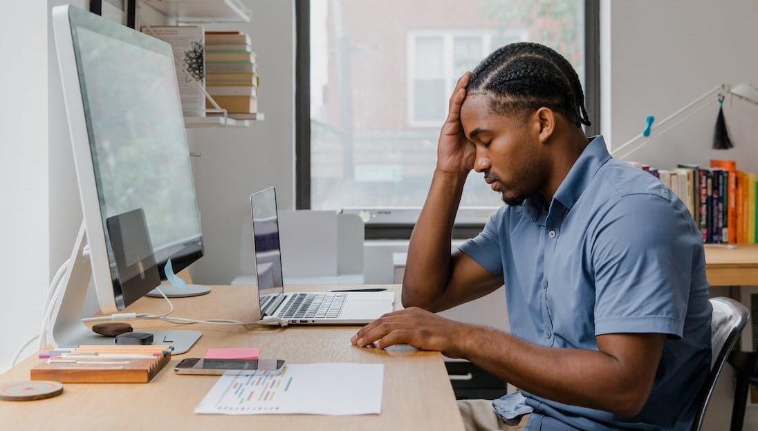 A person sits at a home office desk with their head in their hand, staring downward.