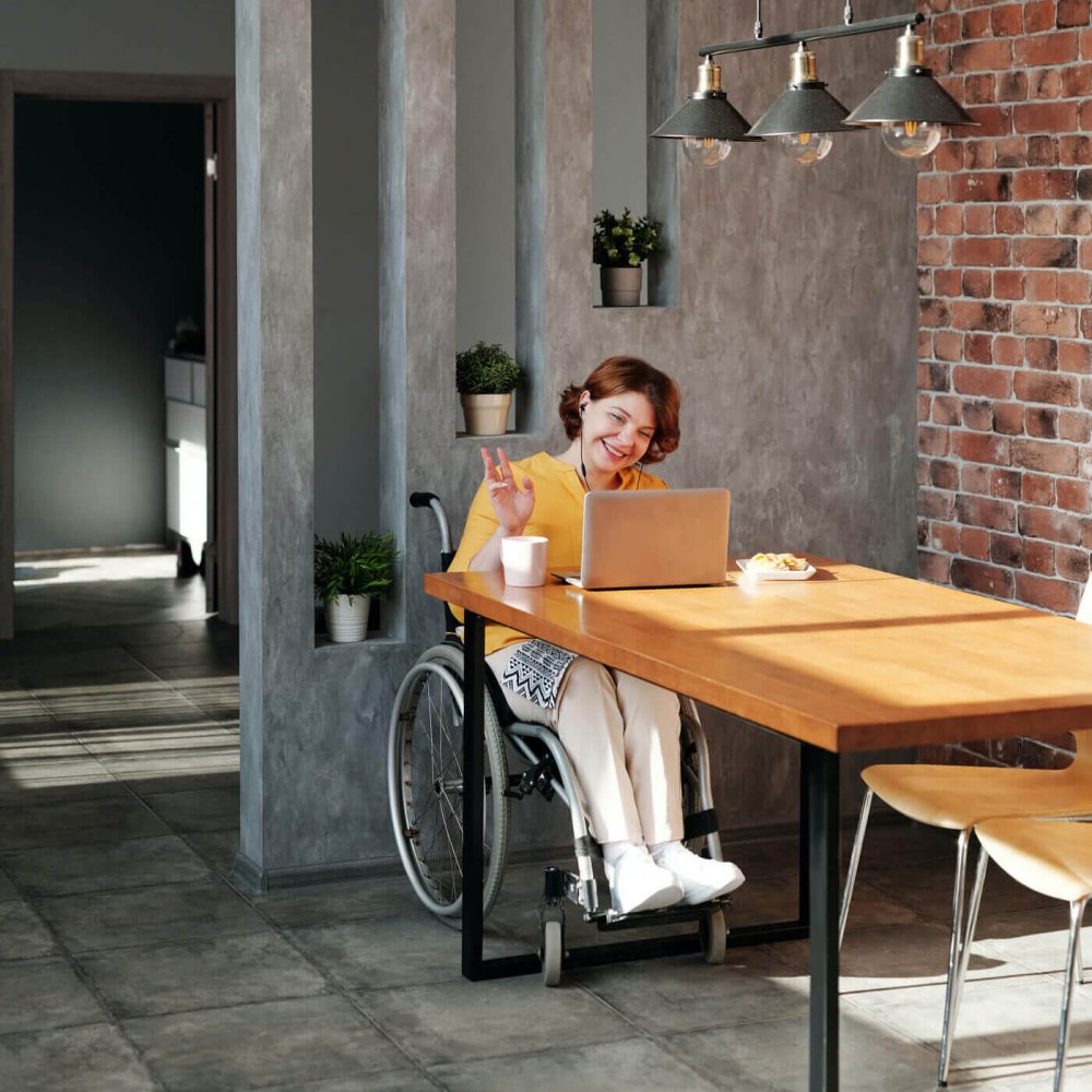 A young professional sitting in a wheelchair waves at the web-camera on her laptop